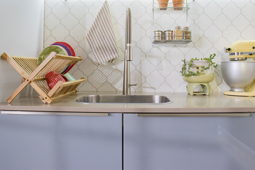 Cozinha com pia de aço inox, utensílios de prato à esquerda, uma panela decorativa com plantas, uma batedeira amarela e uma prateleira com temperos na parede.