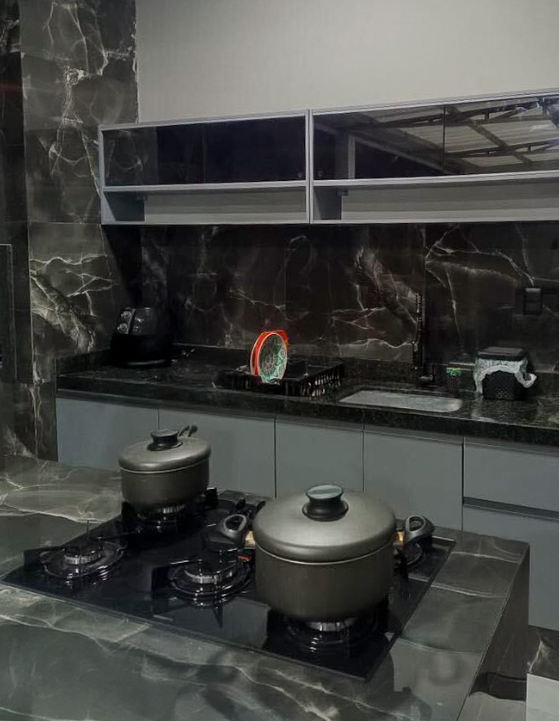 Uma cozinha com bancada preta, armários de aparência moderna e uma área de preparo com panelas, utensílios e alimentos ao fundo