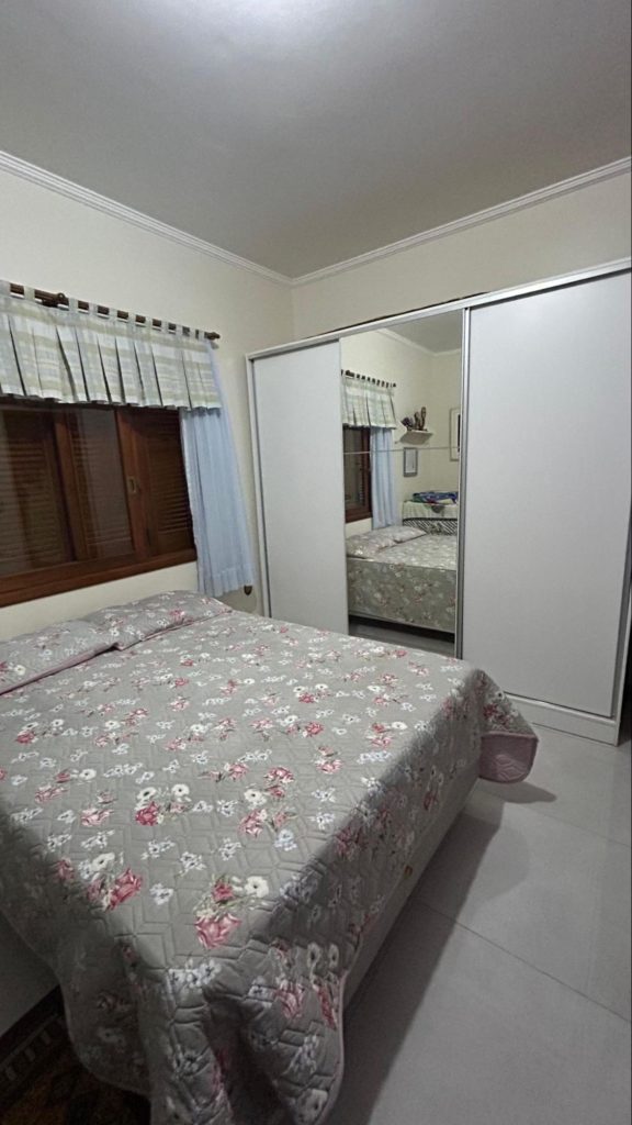 Guarda-roupa com porta espelhada. Ao lado, uma cama de casal com colcha florida abaixo de uma janela de madeira.