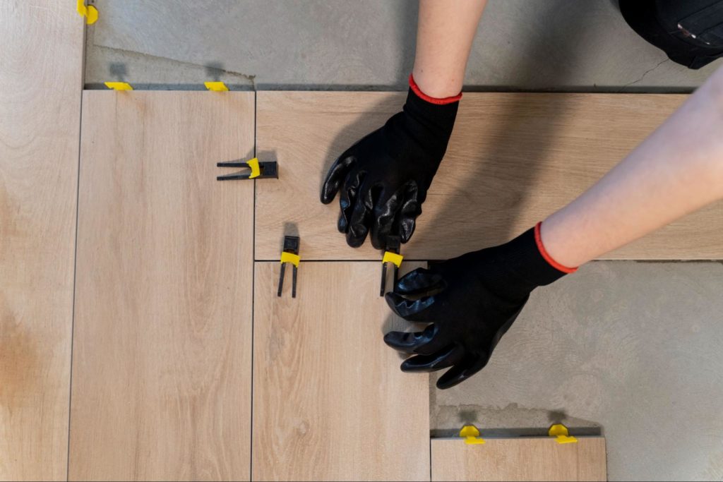 Mãos de uma pessoa usando luvas pretas estão encaixando um piso de madeira em uma instalação. Algumas peças de piso já estão colocadas, e várias presilhas amarelas estão fixando as peças no lugar.
