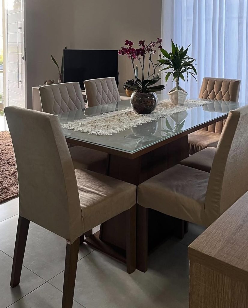 Sala de jantar com mesa de vidro retangular, cercada por seis cadeiras estofadas em tom bege, decorada com vasos de plantas ao centro, próxima a uma janela grande com cortinas brancas.