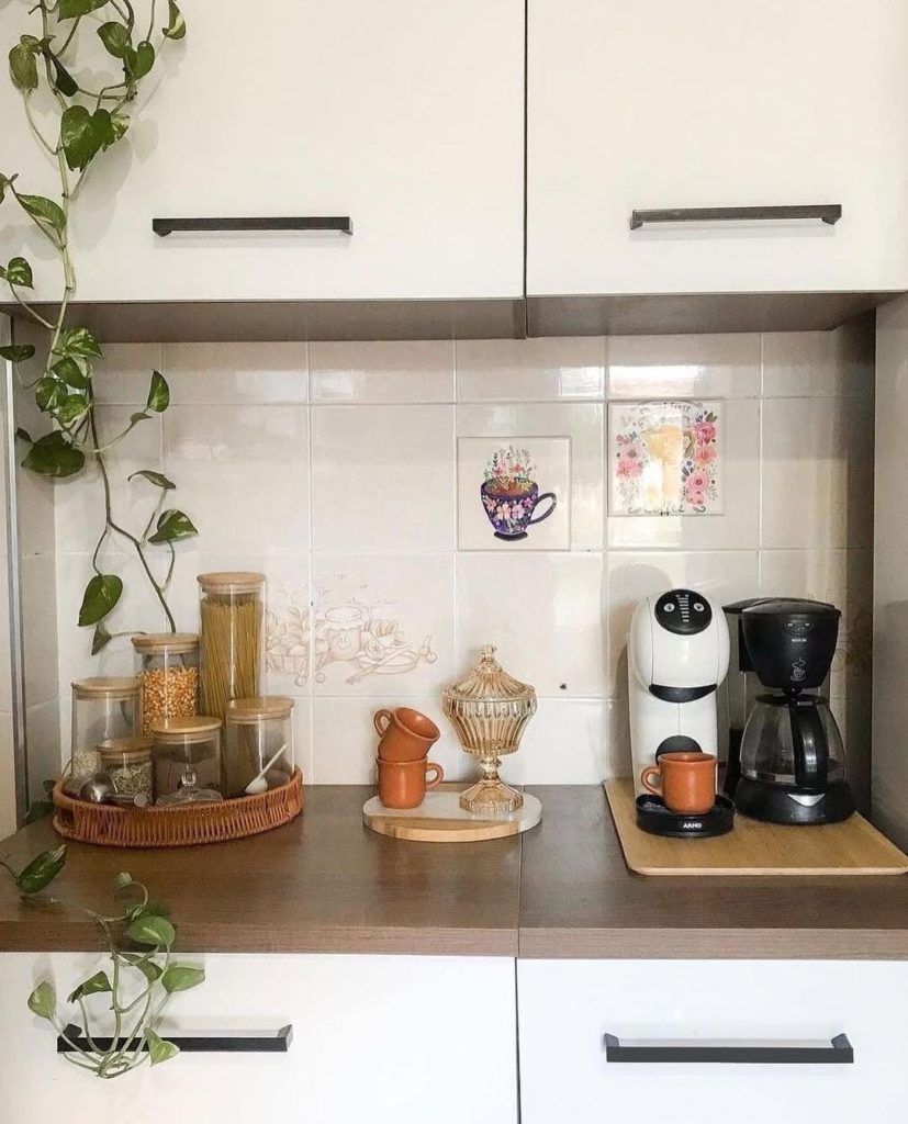 Bancada de cozinha com um cantinho do café organizado, potes de mantimentos em uma bandeja de vime, uma planta pendente e armários brancos ao fundo.