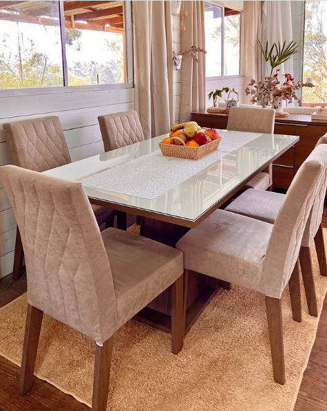 Mesa de jantar retangular com tampo de vidro, cercada por seis cadeiras estofadas em tom bege, com uma cesta de frutas no centro, ao lado de janelas grandes com cortinas claras e plantas decorativas ao fundo.