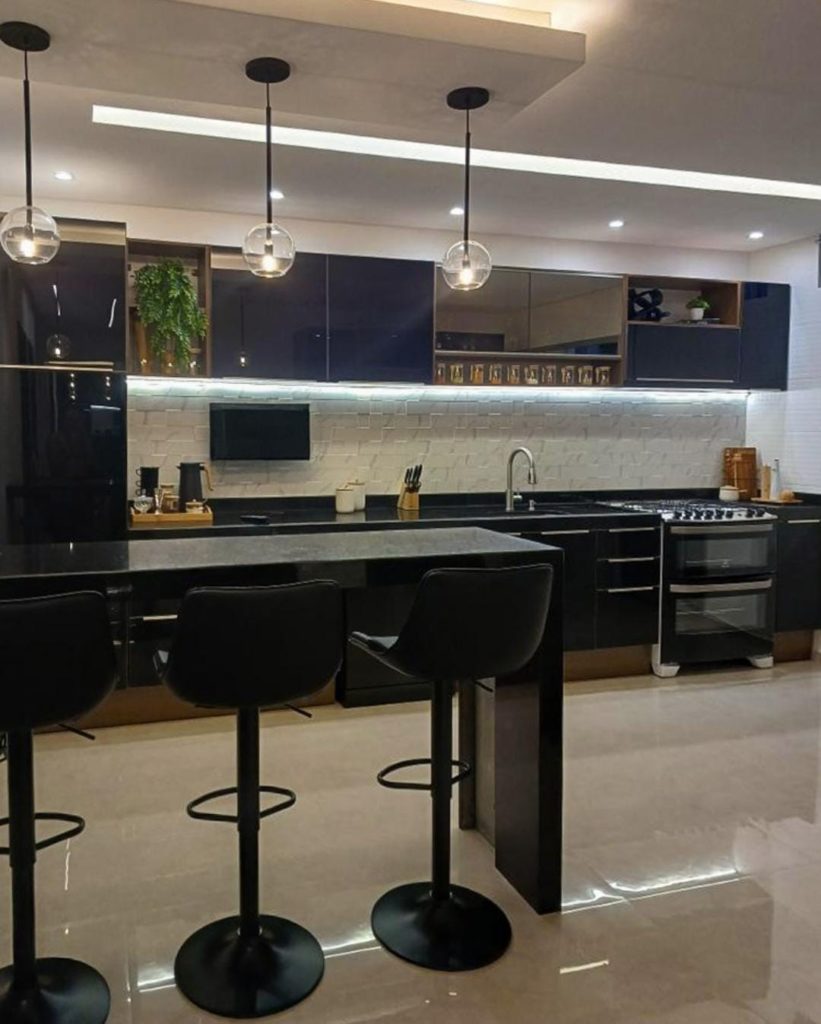 Cozinha com armários pretos, ilha e banquetas pretas. Ao fundo, parede com azulejo branco texturizado e iluminação de LED sob os armários.