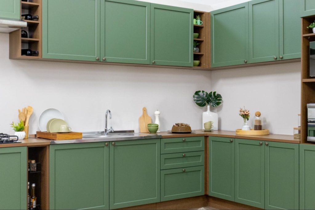 Cozinha com armários verdes, prateleiras de madeira com utensílios e plantas, pia com torneira, e bancada decorada com vasos, pratos, bolo e objetos de cozinha.