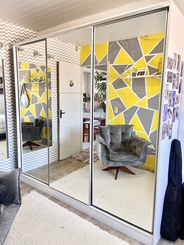 Guarda-roupa com portas de espelho refletindo um quarto com parede geométrica amarela e cinza e uma poltrona de veludo.