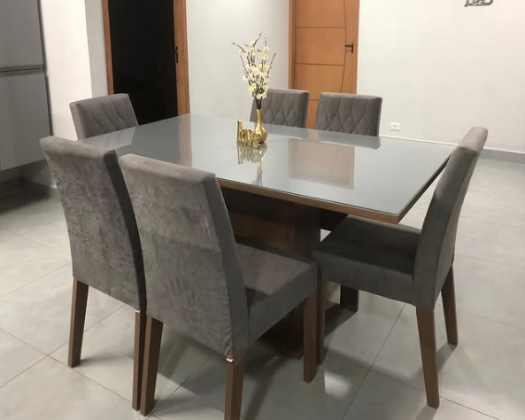 Mesa de jantar retangular com tampo de vidro e seis cadeiras cinza, decorada com um vaso dourado com flores brancas, em um ambiente com piso claro e paredes neutras.