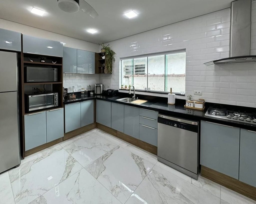 Cozinha moderna com armários cinzas, bancada preta, pia sob uma grande janela, paredes revestidas de azulejos brancos e piso de porcelanato.