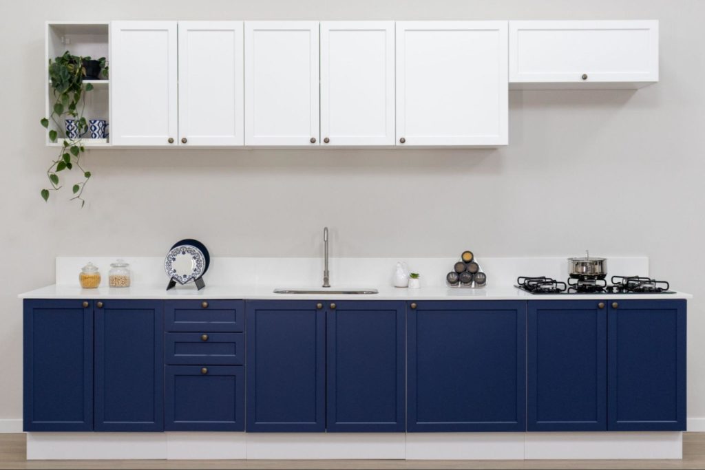Balcão de cozinha com armários azuis na parte inferior, com três gavetas ao centro, e armários brancos na parte superior. No balcão, há pratos decorativos, utensílios de cozinha, uma pia com torneira, e uma panela no fogão ao lado.