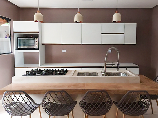 Cozinha com bancada de mármore branco e ilha de madeira, equipada com pia e cooktop. Há armários brancos acima da bancada, um micro-ondas embutido na parede e três pendentes brancos com detalhes em cobre. Quatro cadeiras de design moderno estão posicionadas na ilha.