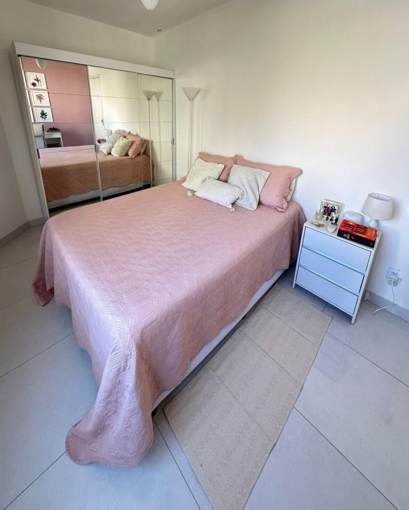 Quarto com cama de edredom rosa-claro, almofadas, cômoda branca com objetos, luminária e guarda-roupa com portas espelhadas refletindo a cama e o ambiente.