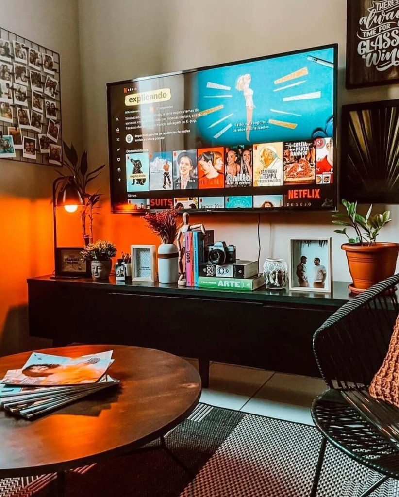 Sala de estar aconchegante com uma TV na parede, exibindo a tela da Netflix. Abaixo, um rack preto está repleto de decorações como livros, uma câmera vintage e porta-retratos. A parede atrás da TV é iluminada por uma luz laranja quente. Ao lado, há um mural de fotos e, no primeiro plano, uma mesa de centro e uma cadeira preta.
