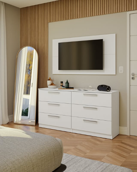 Quarto com uma cômoda branca de seis gavetas, uma televisão na parede acima e um espelho de corpo inteiro apoiado na parede ao lado. Sobre a cômoda, há objetos decorativos e eletrônicos.