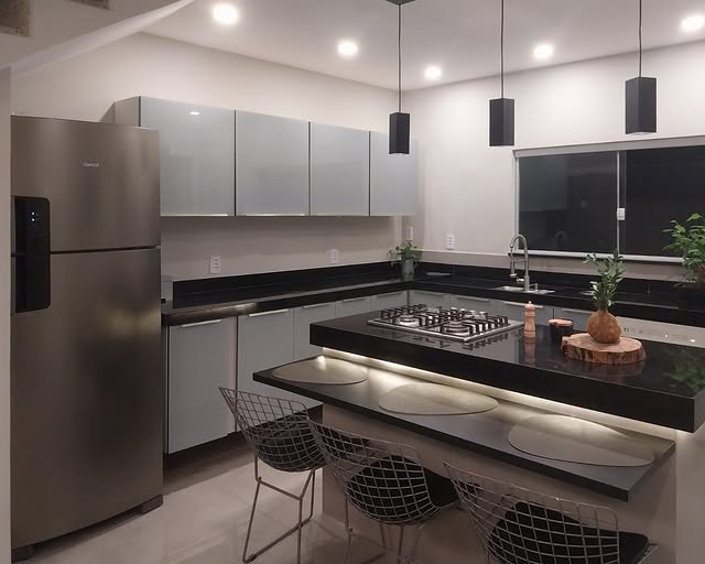 Cozinha com armários cinzas, bancadas de granito preto e uma ilha central com cooktop. A ilha possui uma bancada mais baixa para refeições, iluminada por uma fita de LED e acompanhada por três cadeiras de design aramado. Três pendentes pretos e uma geladeira de inox completam o ambiente.