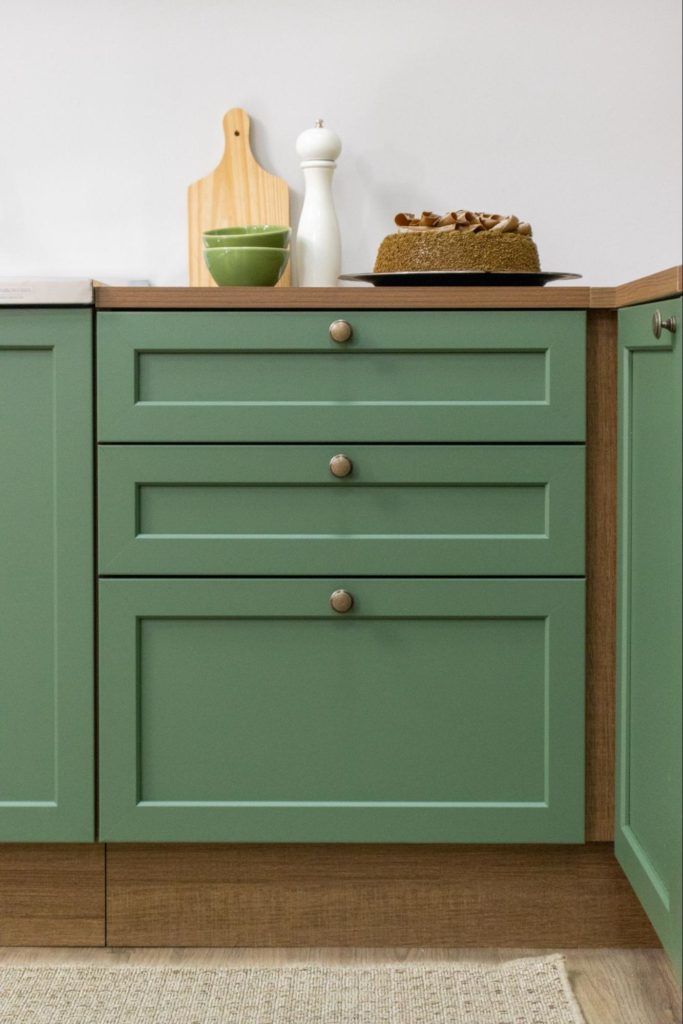 Parte de um armário de cozinha verde com três gavetas, com detalhes em botões metálicos, e uma bancada de madeira clara. No topo, há uma tábua de cortar de madeira, dois bowls verdes, um saleiro branco, um bolo decorado e uma panela no fogão ao lado.