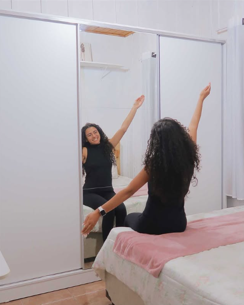 Uma mulher com cabelos cacheados, vestindo roupa preta, sentada na cama de frente para um espelho grande, com os braços levantados e sorrindo.