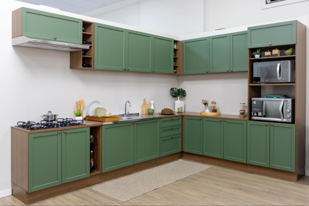 Cozinha com armários verdes na parte inferior e superior, formando um canto. Há uma pia, fogão a gás, micro-ondas, forno de micro-ondas, e utensílios decorativos na bancada.