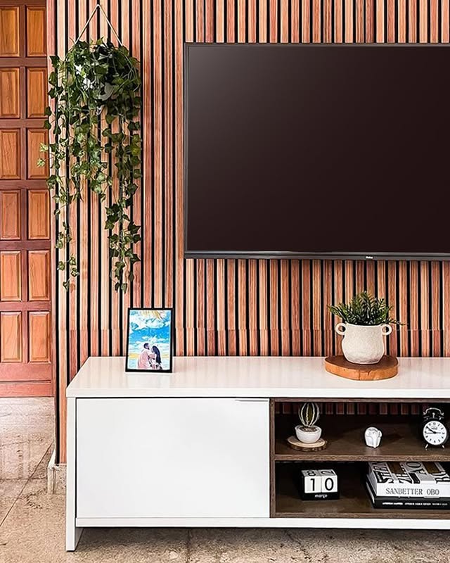 Interior de uma sala com painel de madeira na parede, uma televisão grande pendurada na parede, uma planta pendurada em um suporte na parede, uma planta em um vaso decorativo branco sobre uma bandeja de madeira, uma moldura com uma foto de casal e objetos decorativos na prateleira do móvel.