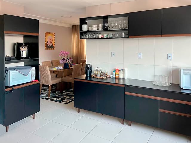Cozinha com armários pretos e detalhes em madeira. Parede de azulejos brancos, um aparador de café ao lado com uma cafeteira, utensílios e objetos decorativos, com uma área de jantar ao fundo com mesa e cadeiras acolchoadas, uma planta de flores na mesa, e uma parede decorada com um retrato.