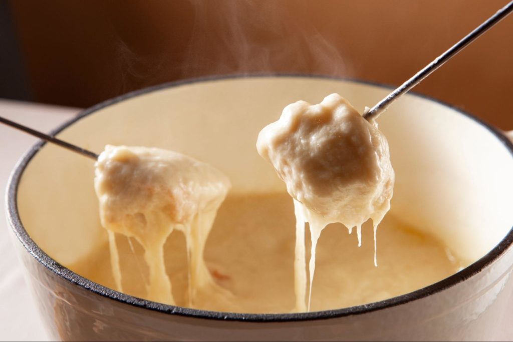 Duas bolas de queijo derretido sendo retiradas de um aparelho de fondue com garfos, com o queijo derretido e borbulhante escorrendo.
