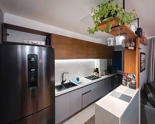 Cozinha com balcão branco na frente, armário aéreo em madeira acima da pia, prateleiras abertas de madeira com plantas, copos e utensílios. Ao lado, uma geladeira inox e uma parede de fundo branca.