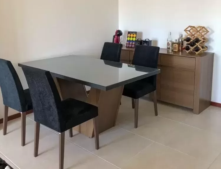 Sala de jantar com mesa retangular de vidro com base de madeira, rodeada por quatro cadeiras estofadas em tecido escuro, e um móvel de buffet de madeira ao fundo, decorado com garrafas de bebidas, uma adega de vinhos, uma máquina de café, e uma bandeja com cápsulas de café.