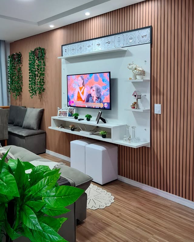 Sala de estar com parede de painéis de madeira, televisão de tela plana instalada na parede, prateleiras brancas decoradas com plantas, objetos decorativos, sofá cinza ao lado, plantas penduradas na parede e uma planta grande no canto inferior esquerdo, além de uma pequena área com tapete de crochê.