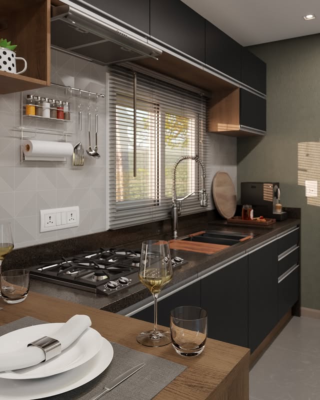 Cozinha com bancadas pretas, janela com persianas, pia com torneira de alta, utensílios pendurados na parede, pratos e copos sobre a mesa de jantar, e uma taça de vinho na frente.