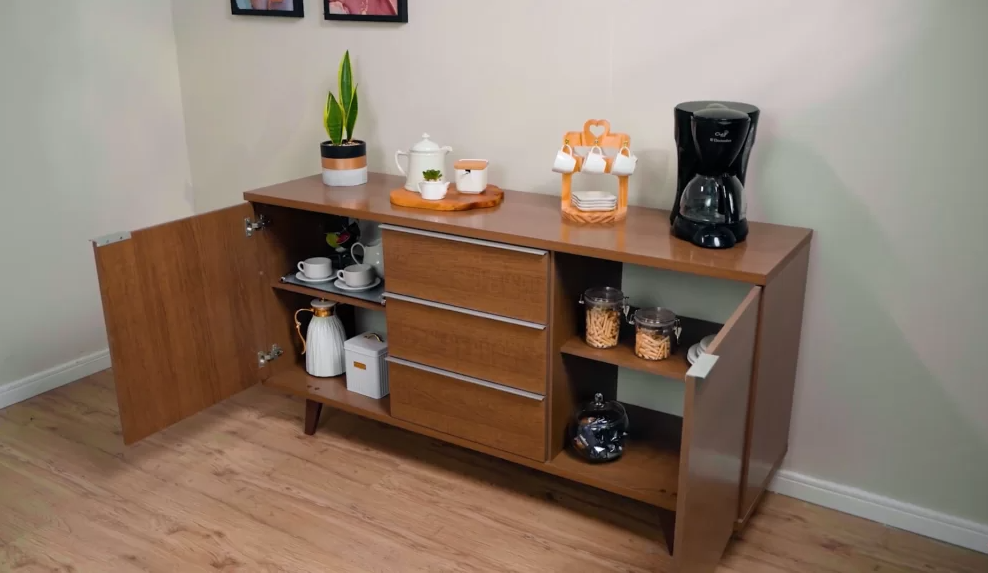 Aparador de madeira com portas abertas e gavetas, colocado contra uma parede branca, com itens de café da manhã e utensílios, incluindo uma cafeteira, jars com cereais, uma garrafa de leite, xícaras, pratos e uma bandeja com objetos decorativos, em um ambiente com piso de madeira.