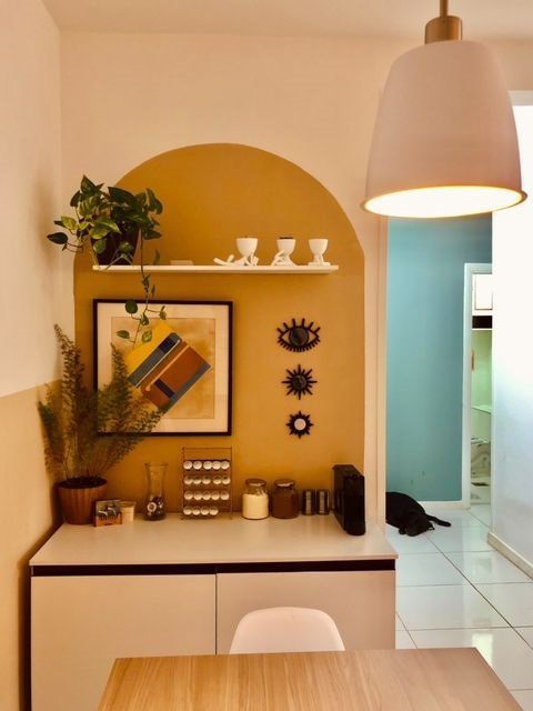 Cantinho decorativo com parede half-tile amarelo, uma prateleira branca com três xícaras brancas e uma planta pendurada, uma obra de arte com formas geométricas, três adornos de sol em metal, uma mesa com vasos, um organizador de cápsulas de café, e pequenos recipientes decorativos. Uma luminária grande branca com detalhes dourados e um pendente amarelo suspenso acima da mesa.