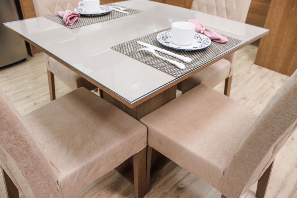 Mesa de jantar quadrada com tampo de vidro, rodeada por quatro cadeiras estofadas em tecido bege. A mesa está posta com jogos americanos, pratos decorativos, xícaras brancas e talheres, com guardanapos cor-de-rosa dobrados ao lado dos pratos.