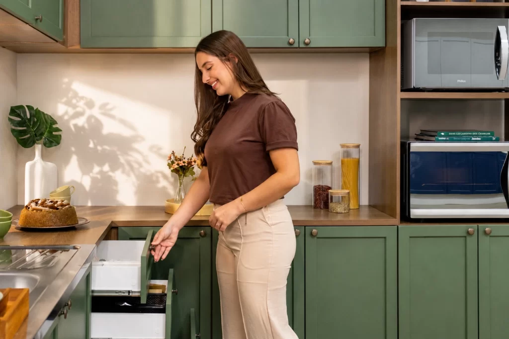 Mulher abrindo uma gaveta com organizadores em uma cozinha planejada verde linha Vik da Madesa, com balcão amadeirado.