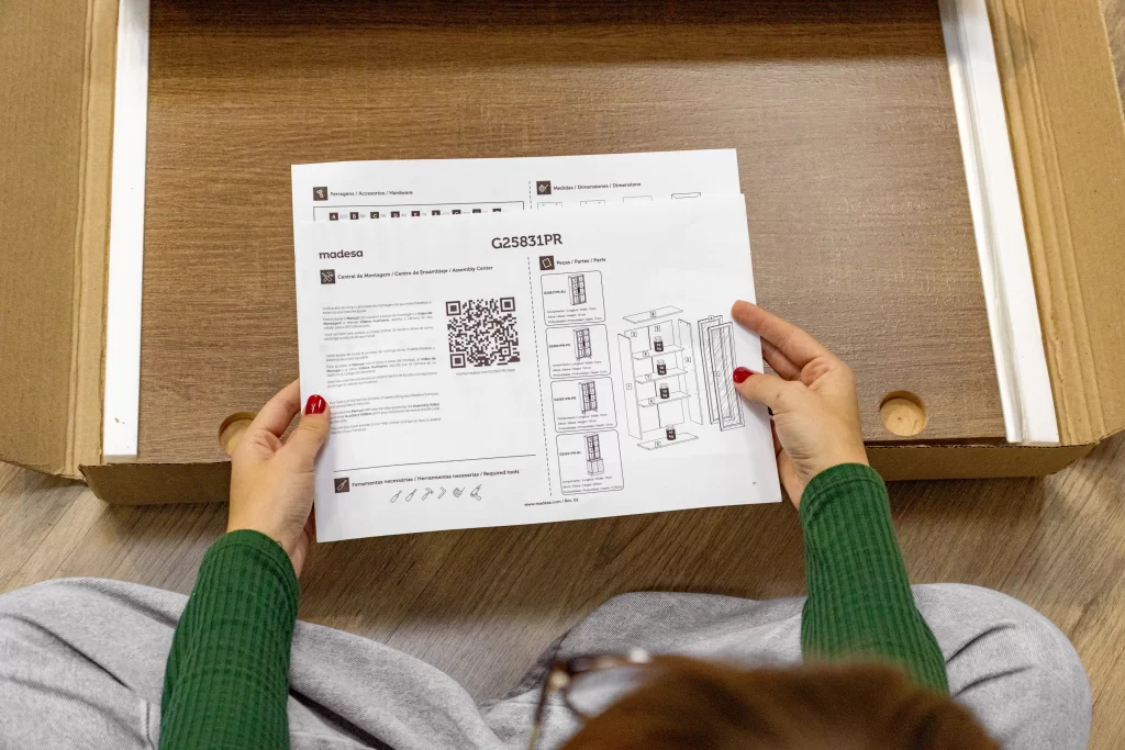 Pessoa segurando o manual de montagem Madesa com ferragens, medidas, peças e QR code, sentada ao lado da caixa aberta