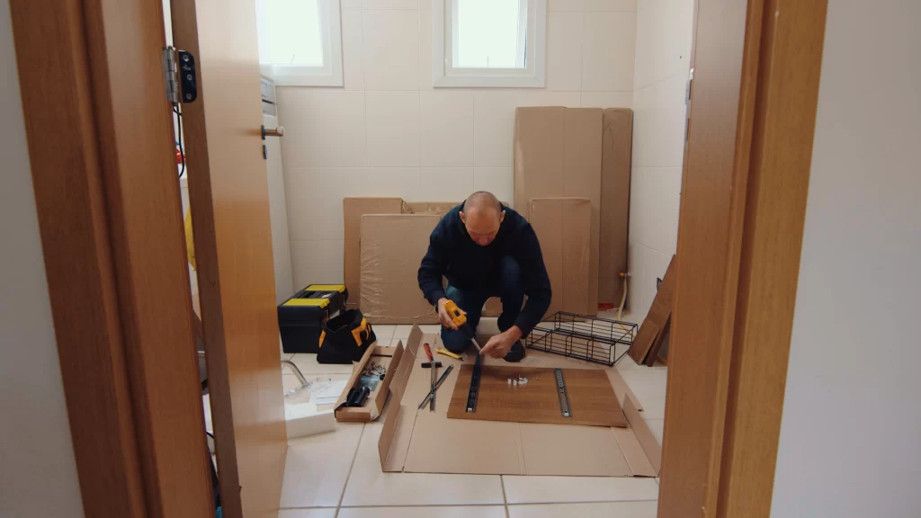 Homem montando móveis em casa com parafusadeira, peças e papelão protegendo o piso, com caixa de ferramentas ao lado