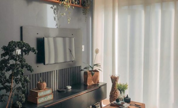 A imagem ilustra uma sala de tv decorada lindamente com itens decorativos como plantas, um baú no centro e uma prateleira de madeira cima da tv.