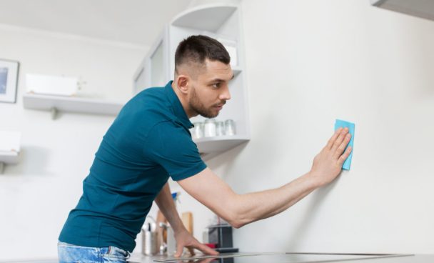 : um homem está limpando a parede da cozinha com um pano azul. Ele usa uma camiseta azul e está concentrado na tarefa, com a mão apoiada na superfície da cozinha. Ao fundo, há prateleiras com utensílios e copos.
