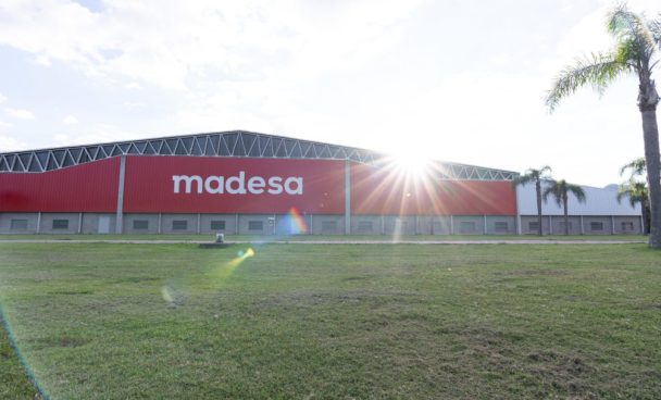 Fábrica vermelha Madesa com céu claro, a frente uma árvore e o gramado