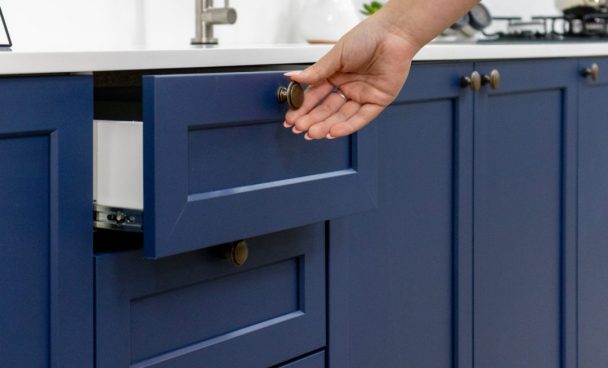 Cozinha Vik na cor azul com caixaria branca. O foco está nas gavetas do balcão de pia sendo abertas por uma mão feminina. Bancada de granito branca com uma torneira de inox ao fundo.