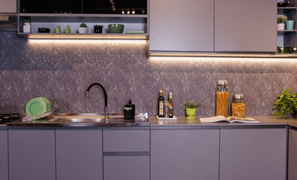 Ambiente com uma pia de cozinha e bancada cinza. A parte superior exibe prateleiras com plantas e utensílios. Louças verdes estão dispostas ao lado da pia.