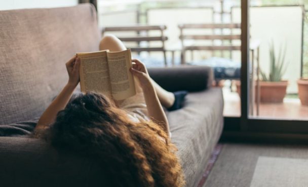 Mulher lendo um livro deitada em um sofá como exemplo de como é morar sozinho