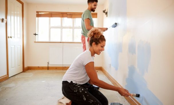 Casal pintando uma parede branca de azul em uma casa que está sendo reformada.