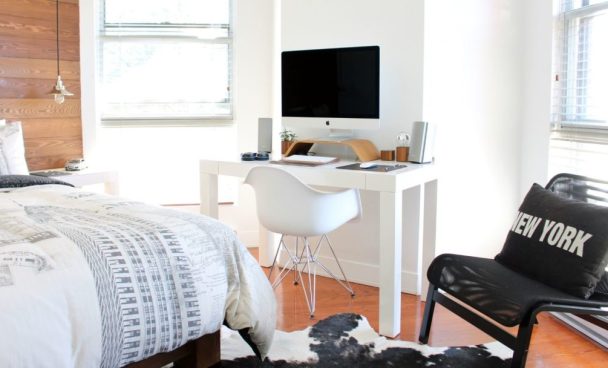 Quarto pequeno em tons claros. Na imagem podemos ver uma parte da cama, uma escrivaninha com computador e uma cadeira com uma almofada preta.