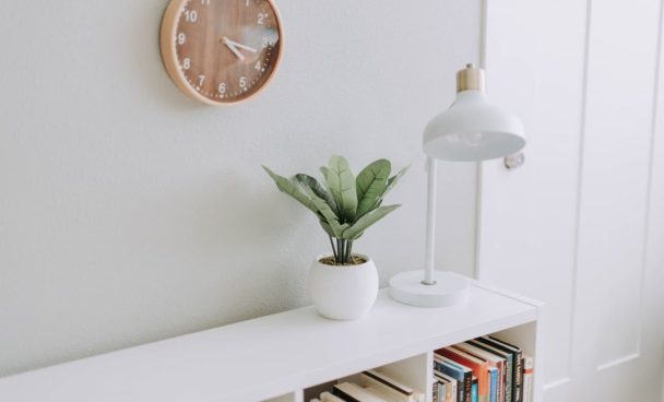 Imagem de uma prateleira de livros brancas decorada como um abajur e um vaso de plantas, itens decorativos que combinam com o minimalismo.
