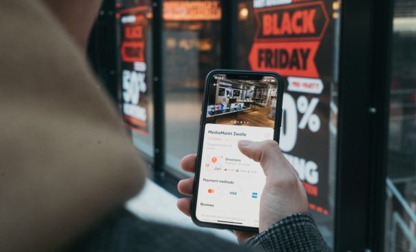 Pessoa conferindo promoções de Black Friday no celular em frente a uma vitrine com descontos anunciados em adesivos vermelhos e brancos.