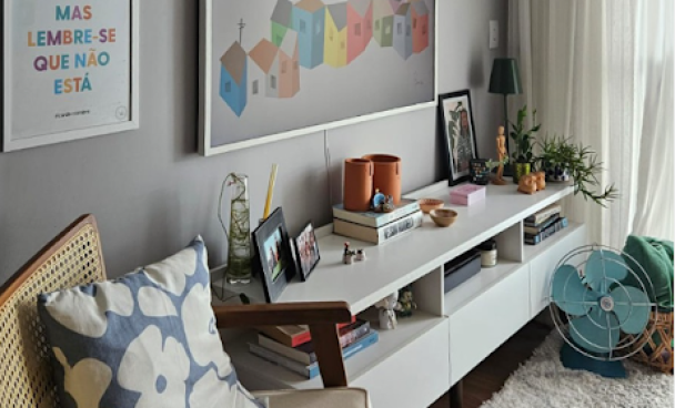 Sala de estar com sofá de madeira e almofada com estampa floral. Uma mesa branca exibe livros, uma caneca, plantas e quadros com fotos.