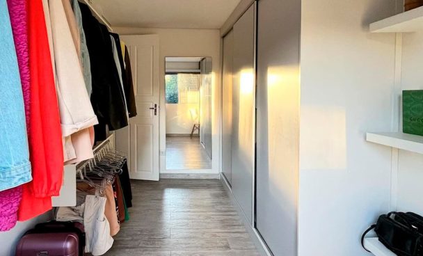 Corredor de quarto com guarda-roupa de correr branco à direita, parede branca, prateleiras brancas com objetos decorativos e uma mala roxa no chão, roupas penduradas em uma parede à esquerda, iluminação natural entrando pelo fundo do corredor, que leva a uma área de varanda ou janela ao fundo.