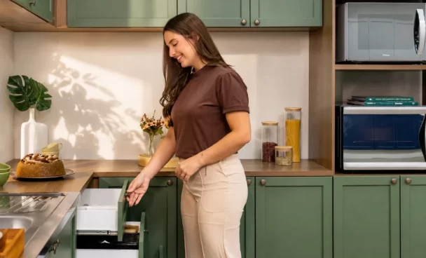 Mulher abrindo uma gaveta com organizadores em uma cozinha planejada verde linha Vik da Madesa, com balcão amadeirado.