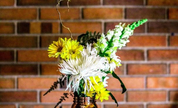 um vaso de metal acobreado com arranjo de flores branca e amarelas, folhagens e galhos secos está em foco, em frente a parede de tijolos à vista