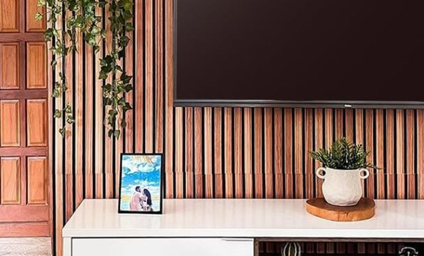 Interior de uma sala com painel de madeira na parede, uma televisão grande pendurada na parede, uma planta pendurada em um suporte na parede, uma planta em um vaso decorativo branco sobre uma bandeja de madeira, uma moldura com uma foto de casal e objetos decorativos na prateleira do móvel.