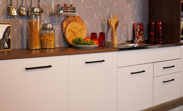 uma cozinha planejada Madesa Ágata na cor branca está em foco. Sobre ela, estão dispostos utensílios de cozinha e alimentos. Na parede do fundo, é possível notar um papel de parede cinza com detalhes em linhas brancas geométricas.